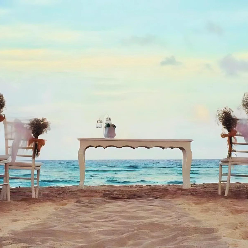 Two chairs flank a white table adorned with lanterns and flowers on a sandy beach, set up for a destination wedding, overlooking a calm sea.