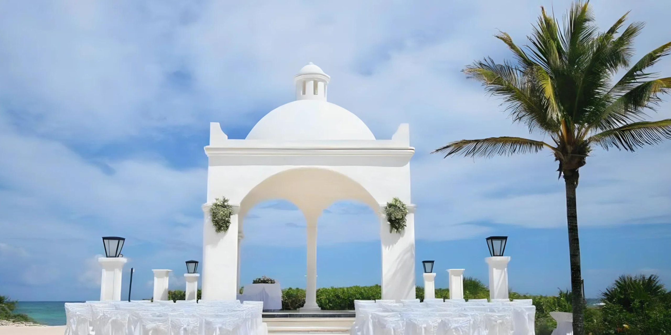 Destination wedding at a white beachfront pavilion with a dome roof, decorated with flowers, flanked by lanterns and palm trees against a blue sky.