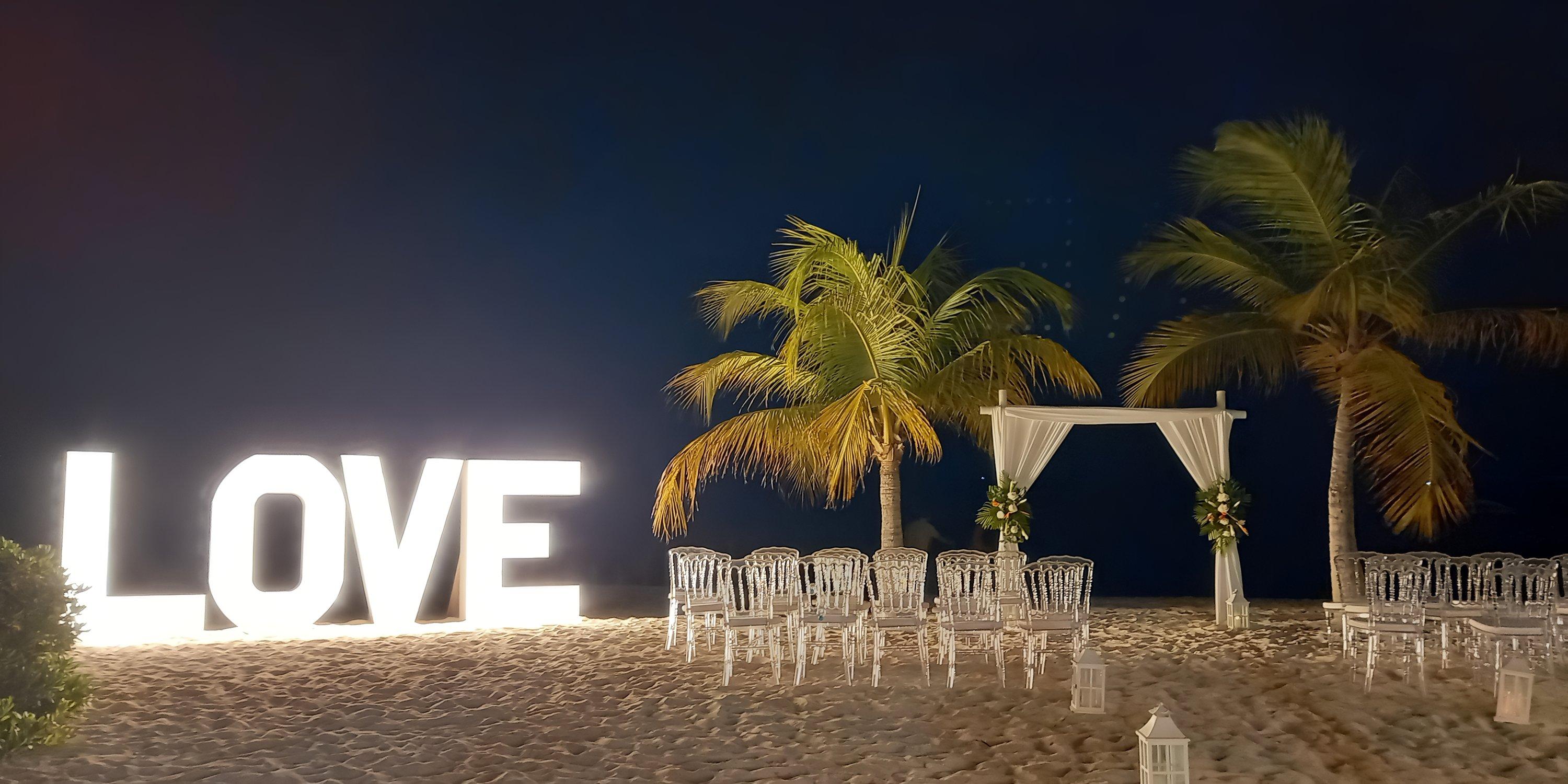 Barcelo Aruba: Destination wedding setup with glowing "LOVE" sign, arch, and chairs on a nighttime beach.