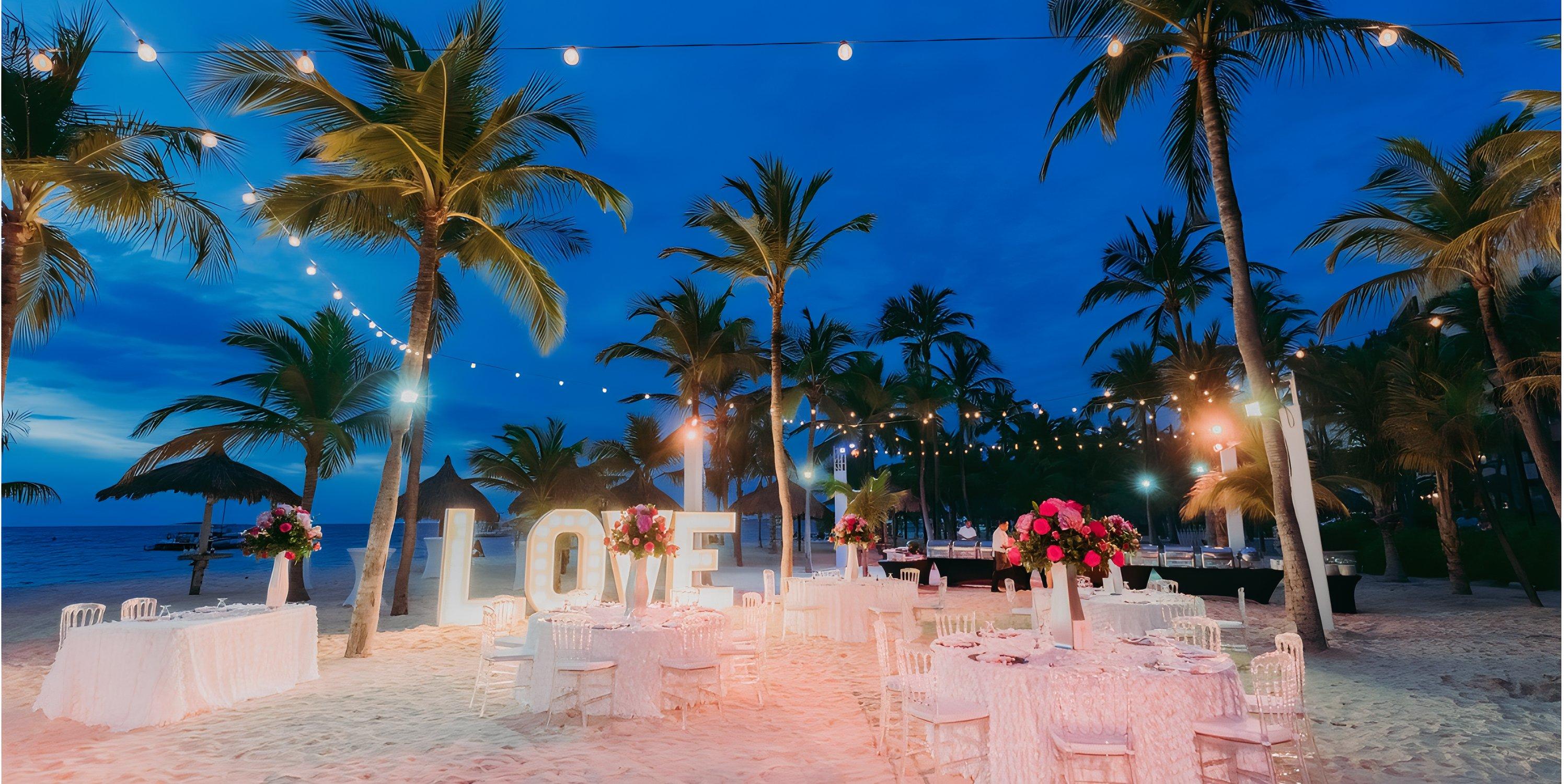 Destination wedding setup at Barcelo Aruba with palm trees, string lights, and large "LOVE" letters at dusk.