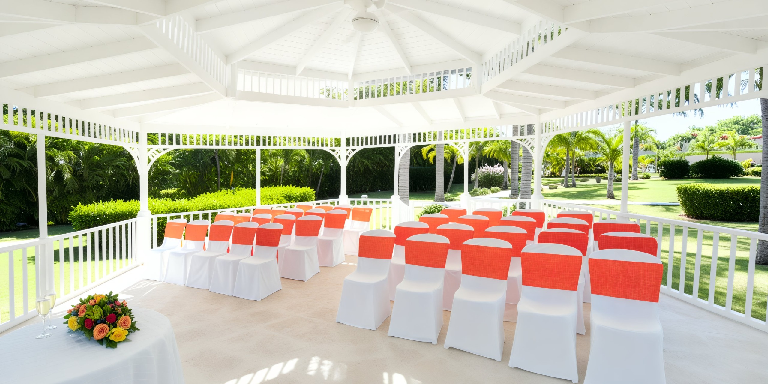 Garden destination wedding featuring a charming outdoor setup with white chairs adorned with vibrant orange sashes, all arranged gracefully under a picturesque gazebo.