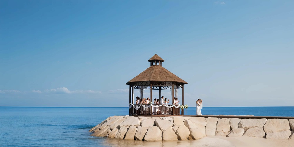 A small wedding ceremony is taking place under a gazebo on a rocky pier by the sea, with a clear blue sky and calm water in the background.