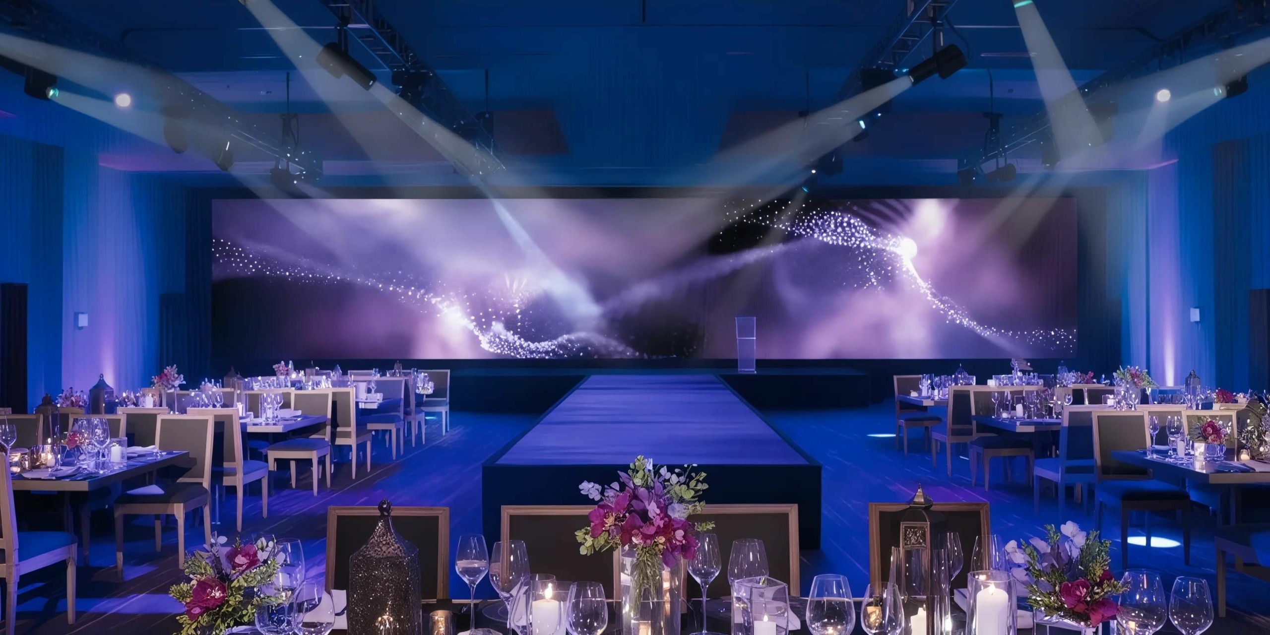 A grand event hall at Secrets Riviera Cancun set up for a formal banquet, featuring an illuminated stage, multiple dining tables adorned with floral centerpieces, and a large screen displaying abstract visuals in the background.
