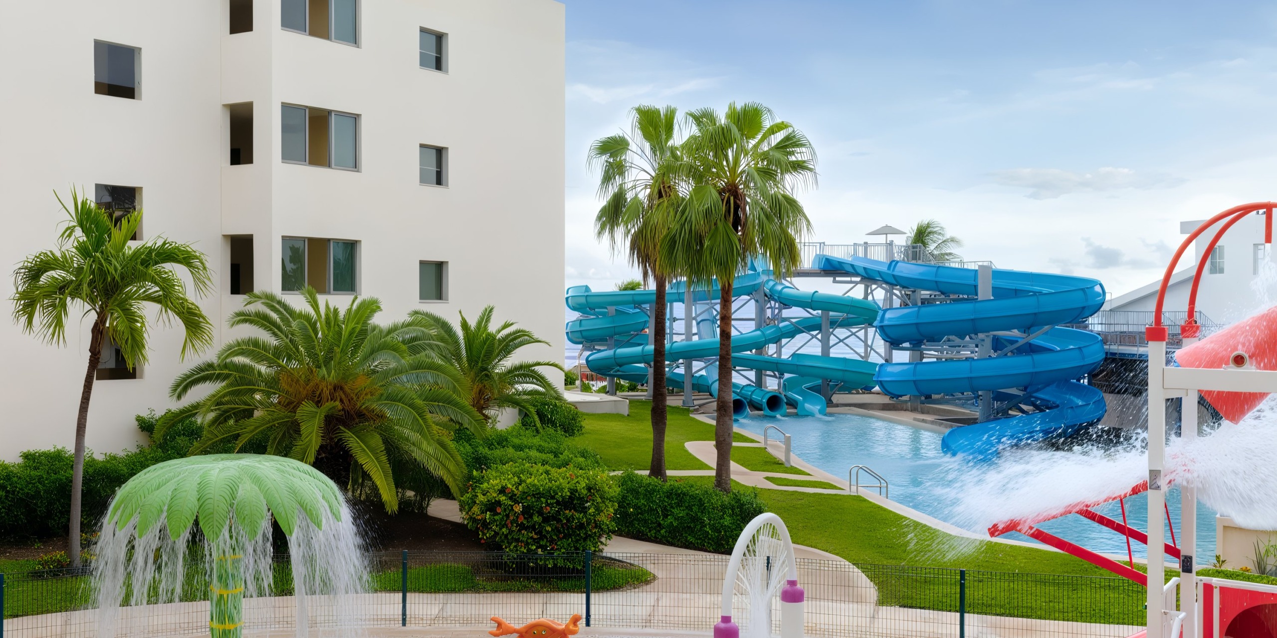 The Hyatt Ziva Riviera Cancun features a resort pool area with expansive blue water slides, palm trees, and a splash pad, complemented by a modern white building situated on the left.