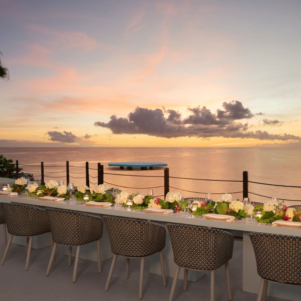 Long table with floral decor set for a destination wedding, overlooking the ocean at sunset and clouds.