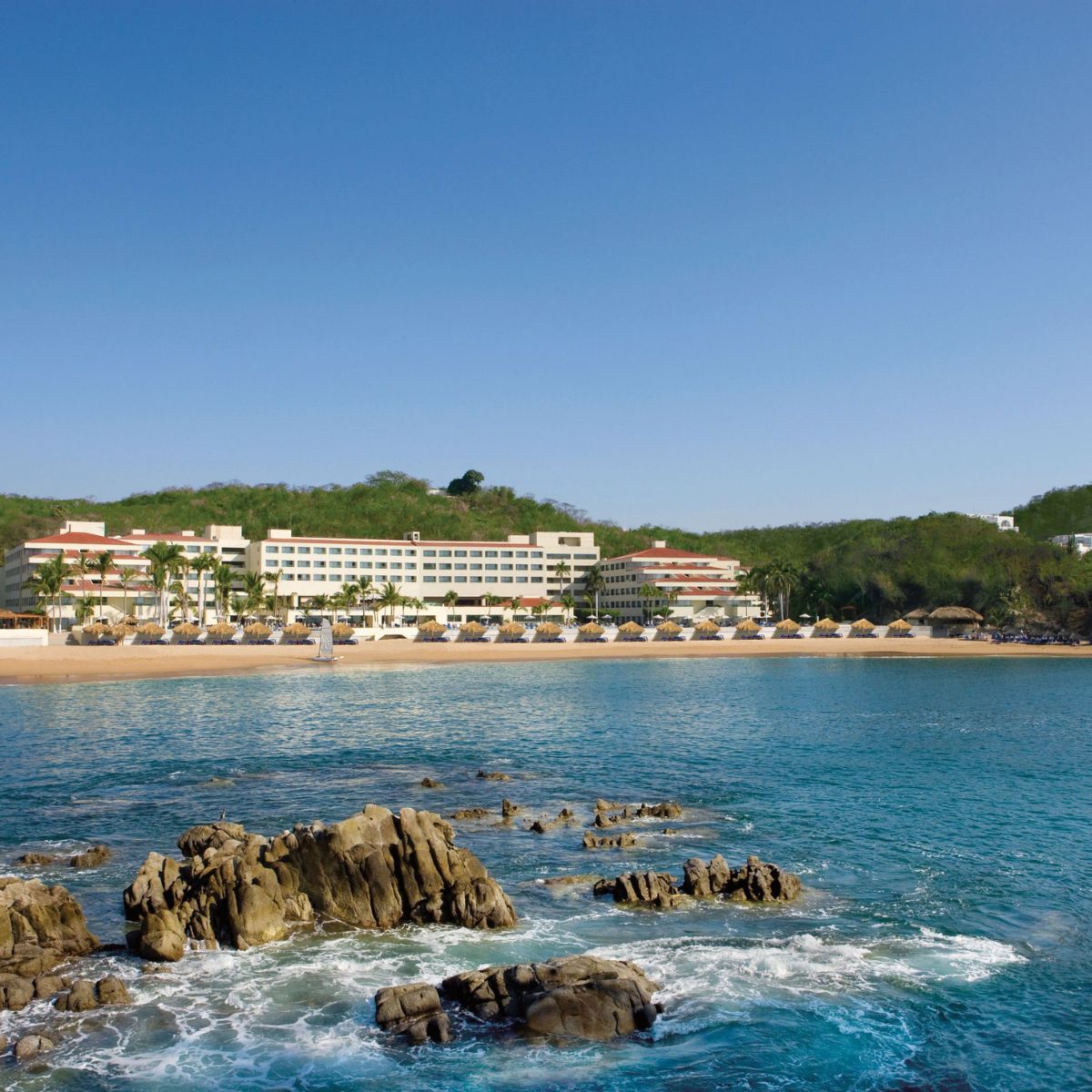 Dreams Huatulco: Destination wedding spot with beachfront hotel, umbrellas, lounge chairs, and rocky ocean.