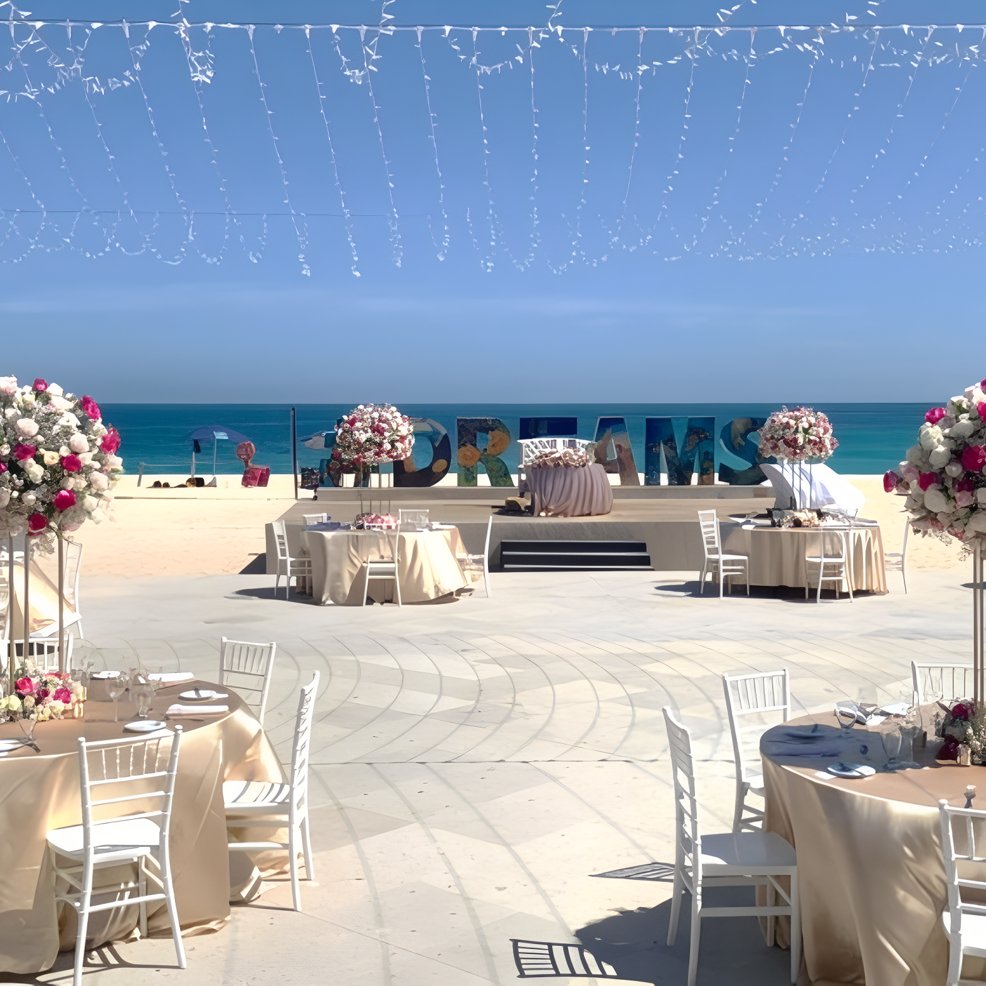 Destination wedding setup on the beach with round tables, floral centerpieces, and "FOR DREAMS" sign.