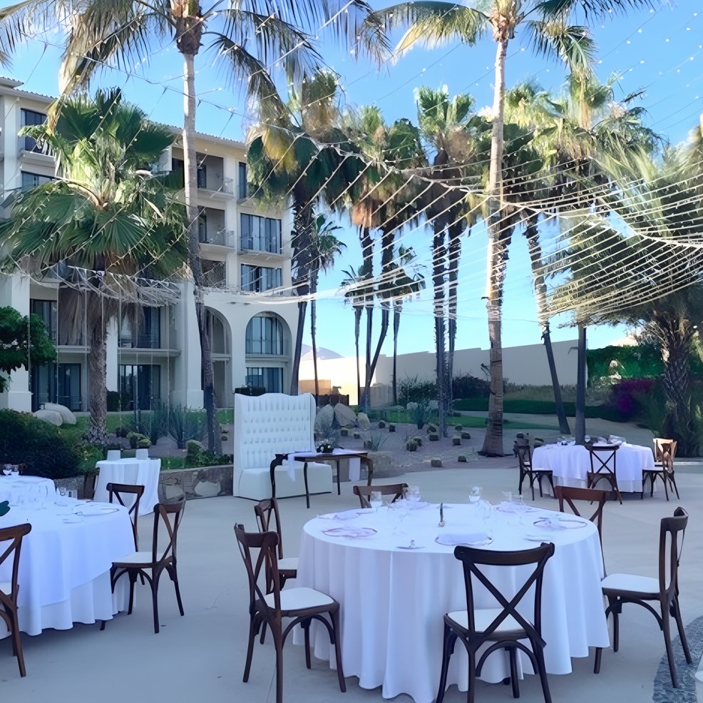 Destination wedding setup featuring round tables draped in white linens and string lights beneath palm trees.