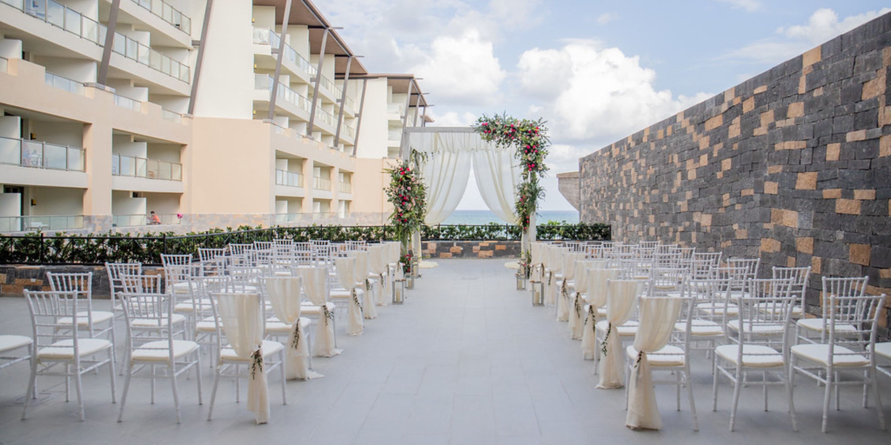 Destination wedding setup at Dreams Natura with white chairs, floral arch, and buildings in the background.