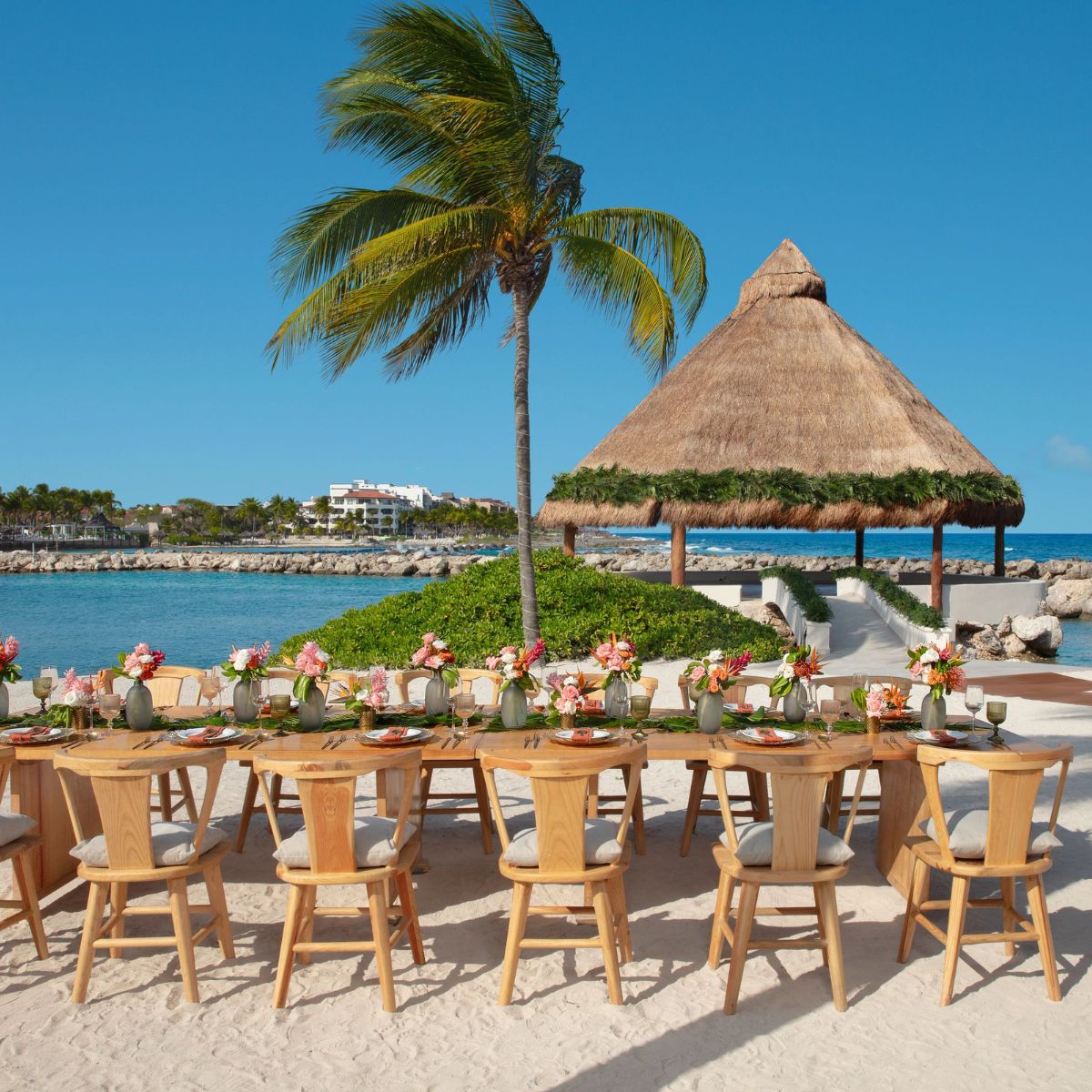 Outdoor dining table with wooden chairs on the beach, ideal for a destination wedding by a gazebo.