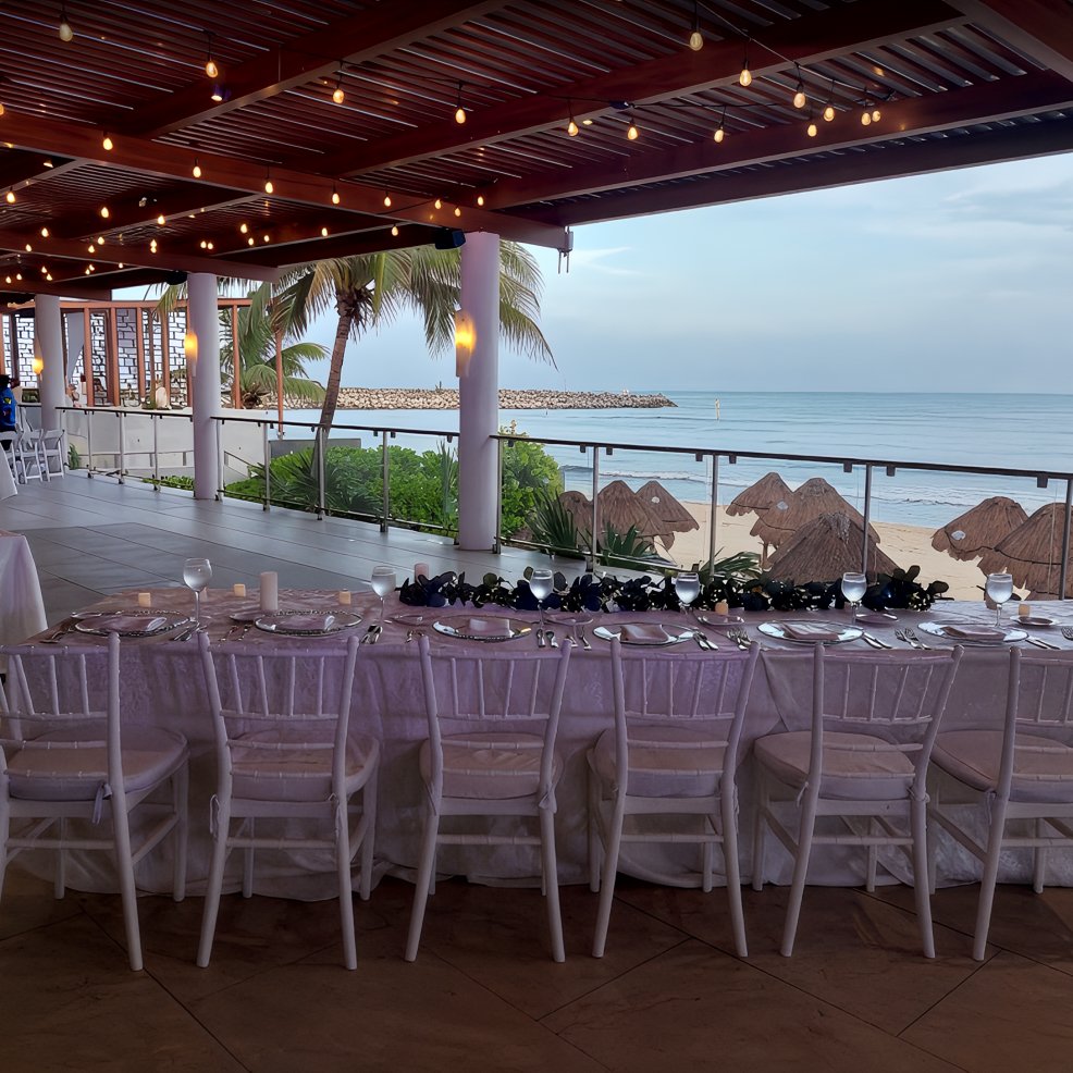 Elegant long table set for a destination wedding on a covered terrace, with views of beach and ocean.
