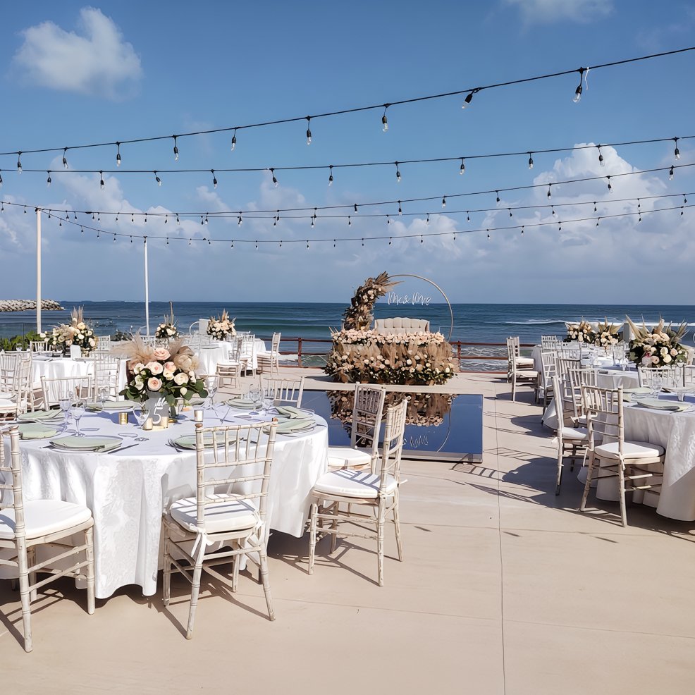 Destination wedding reception outdoors featuring round tables, floral centerpieces, and ocean backdrop.