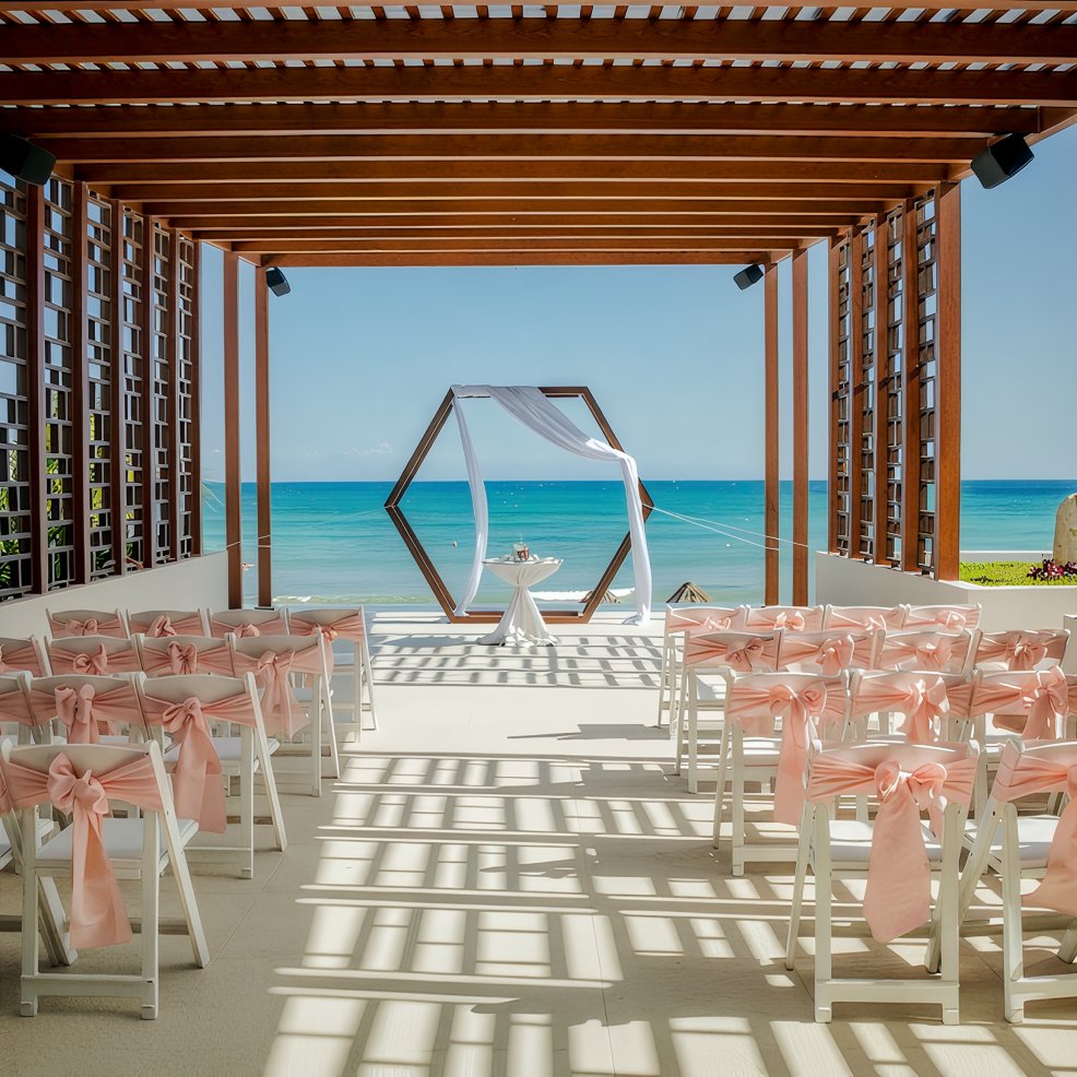Destination wedding ceremony by the sea with pink ribbon chairs and a wooden geometric arch.