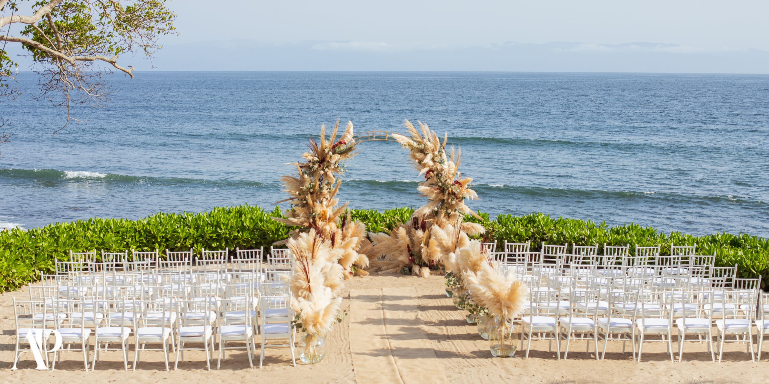 Destination wedding setup at Dreams Bahia Mita with white chairs and a pampas grass arch on the beach.