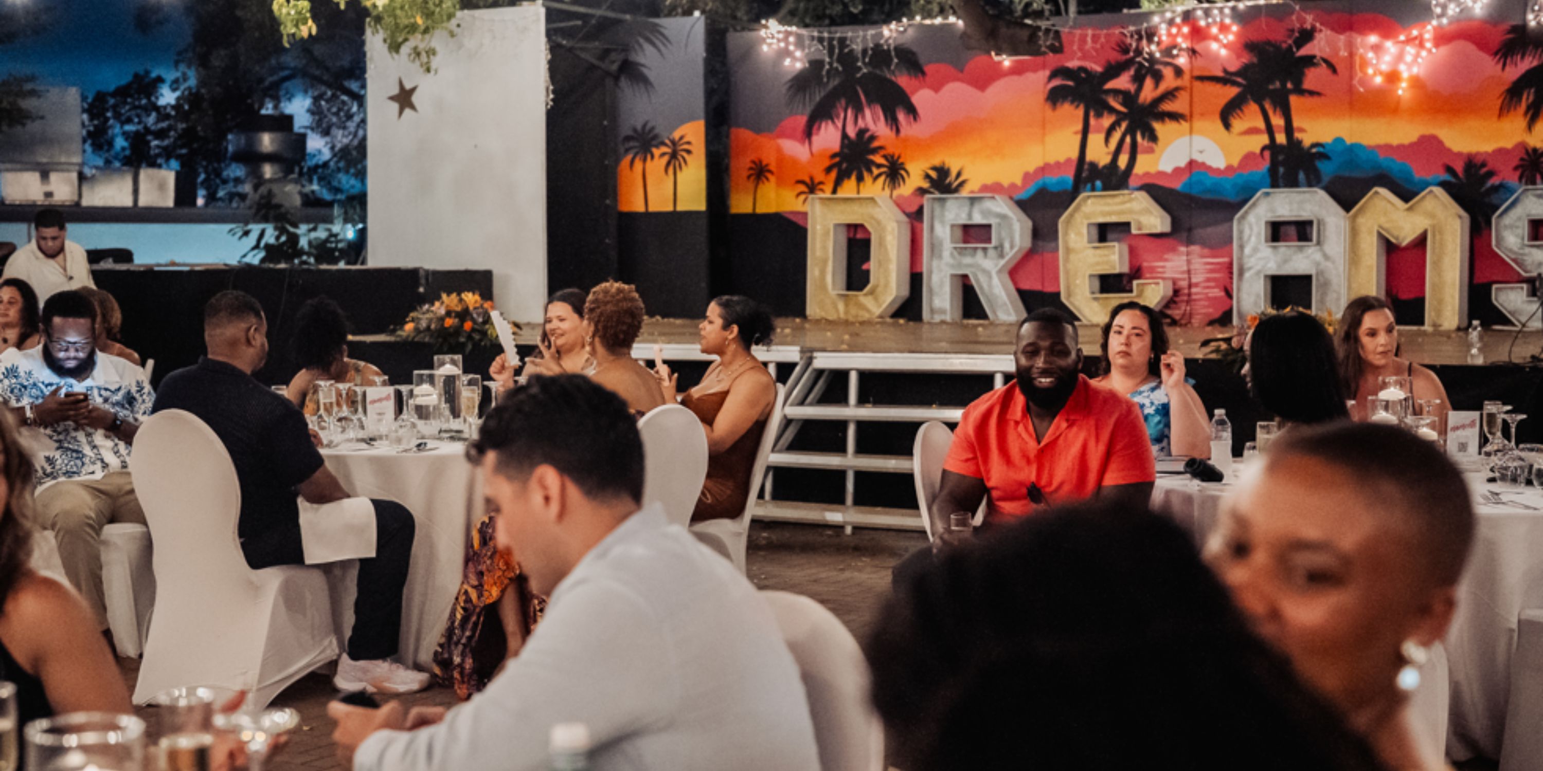 Guests at a destination wedding are seated at decorated tables with a "Dreams Curacao" backdrop.