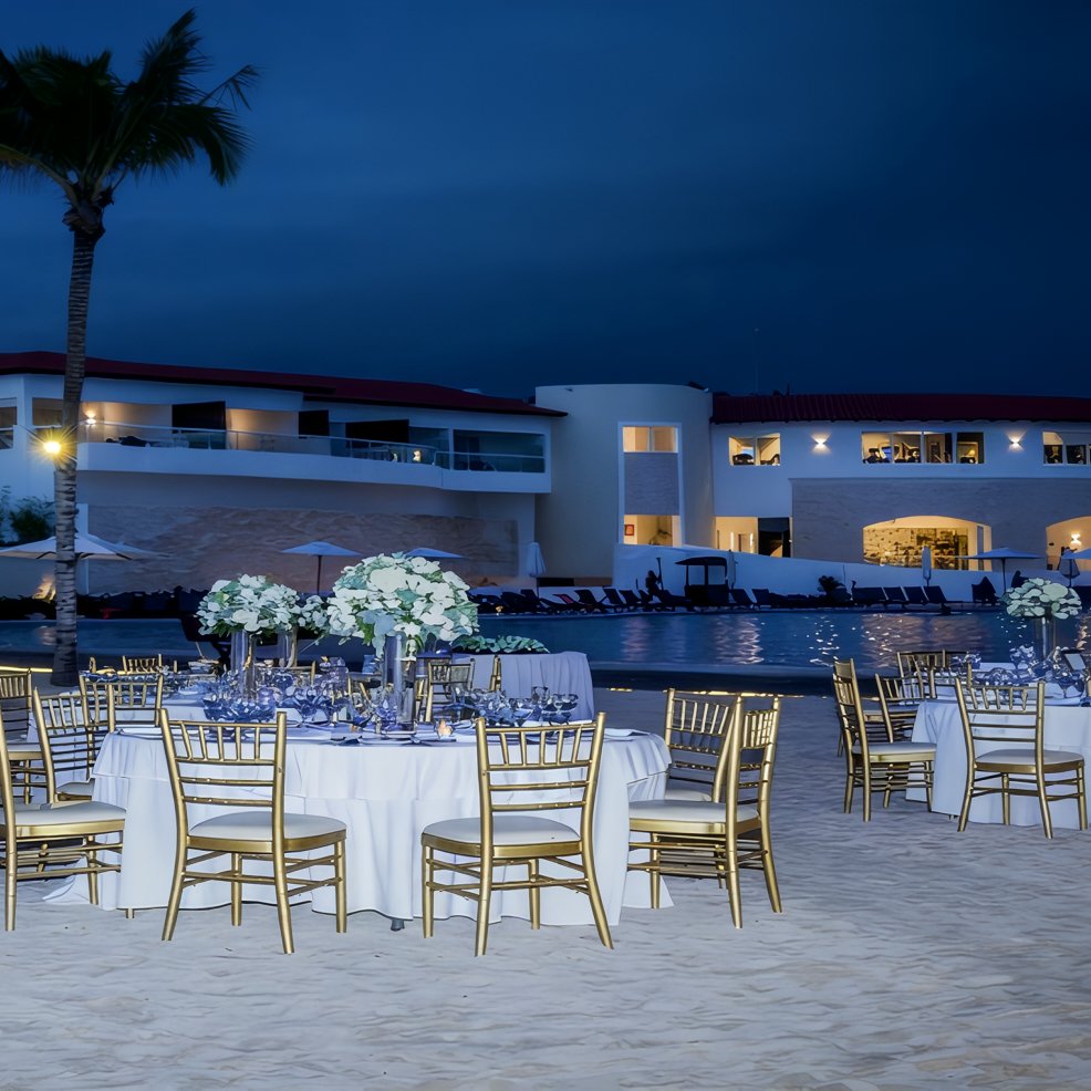 Chic destination wedding dining area with round tables and gold chairs on sand near a glowing resort at night.