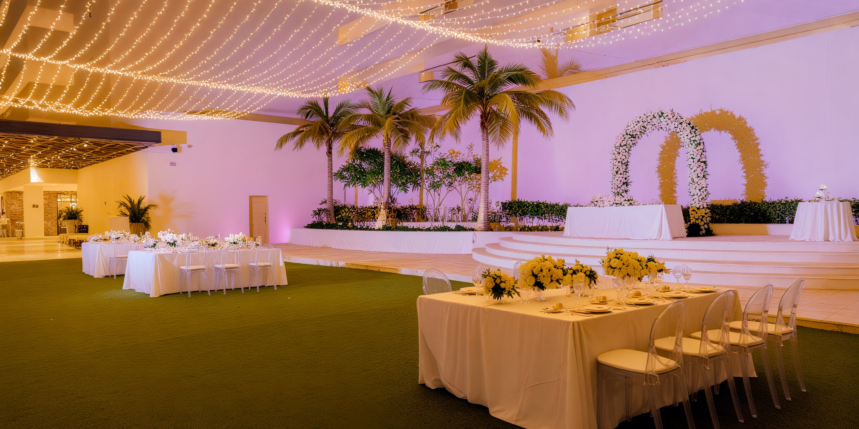 Dreams Natura: An elegant indoor wedding venue featuring beautifully decorated tables, lush palm trees, and a stunning floral arch—perfect for your dream destination wedding.