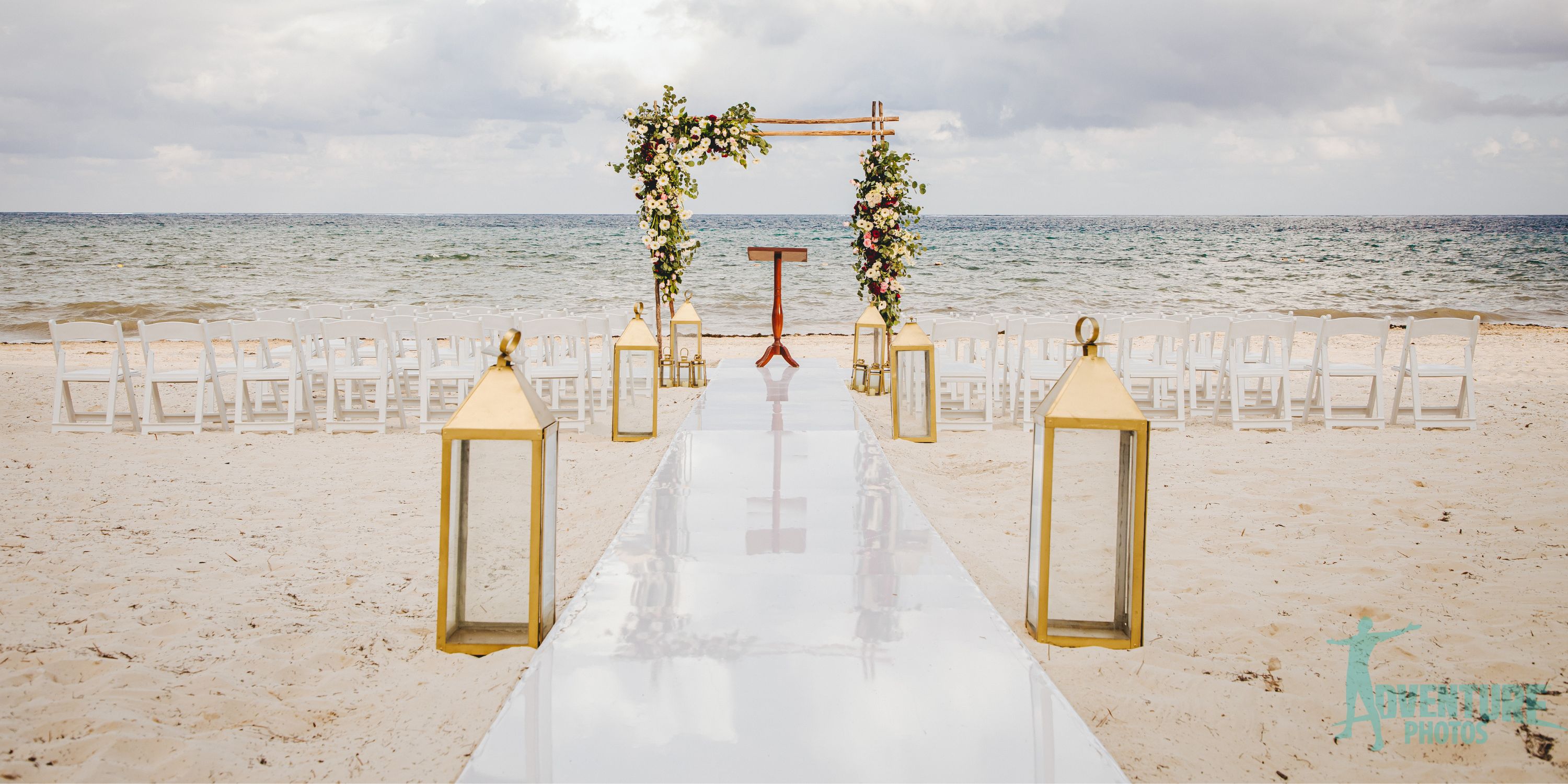 Destination wedding setup at Dreams Sapphire: white chairs, floral arch, gold lanterns by the ocean.