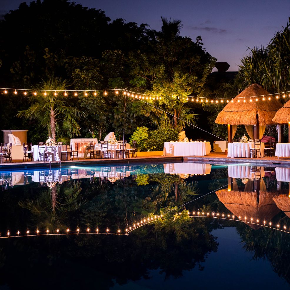 Destination wedding reception by the pool at dusk, with string lights, tables, and thatched huts.