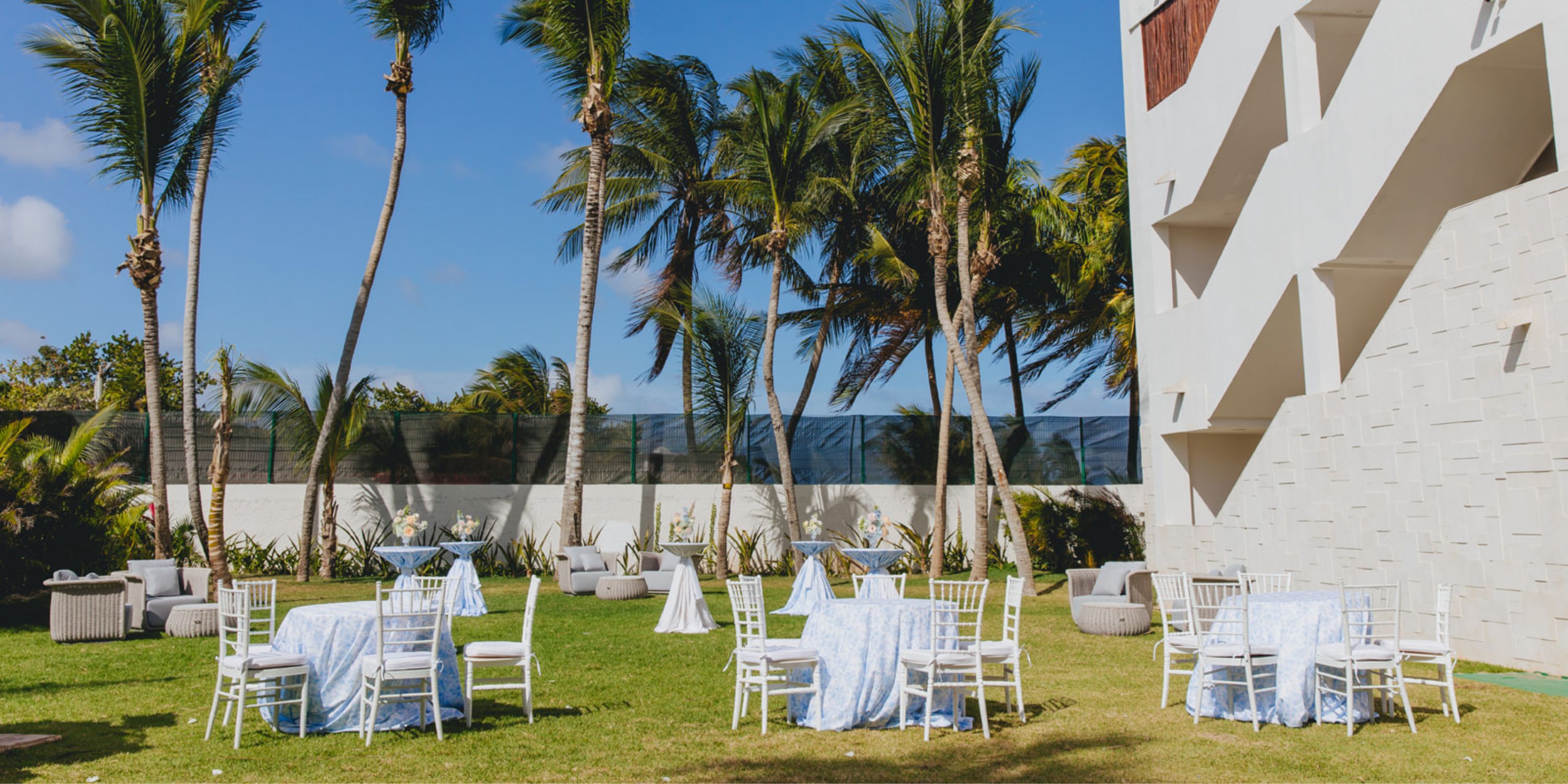 Dreams Sapphire: Elegant outdoor wedding setup with white chairs, tables on grass, palms, modern venue.