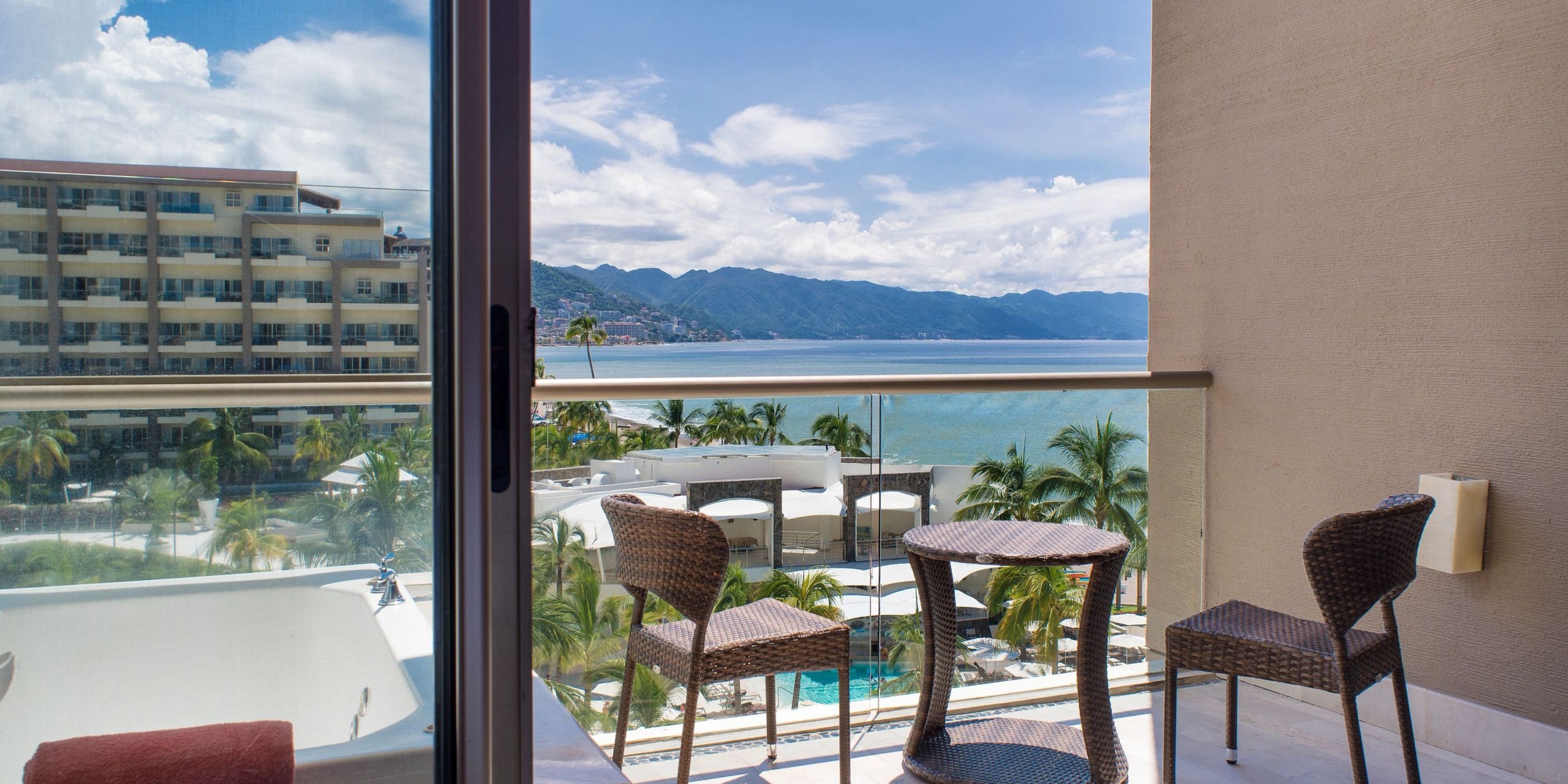 Relax on the balcony of Dreams Vallarta Bay, equipped with two chairs and a table, offering breathtaking views of the resort's swimming pool, lush palm trees, and a stunning coastal vista with mountains in the distance on a clear day.