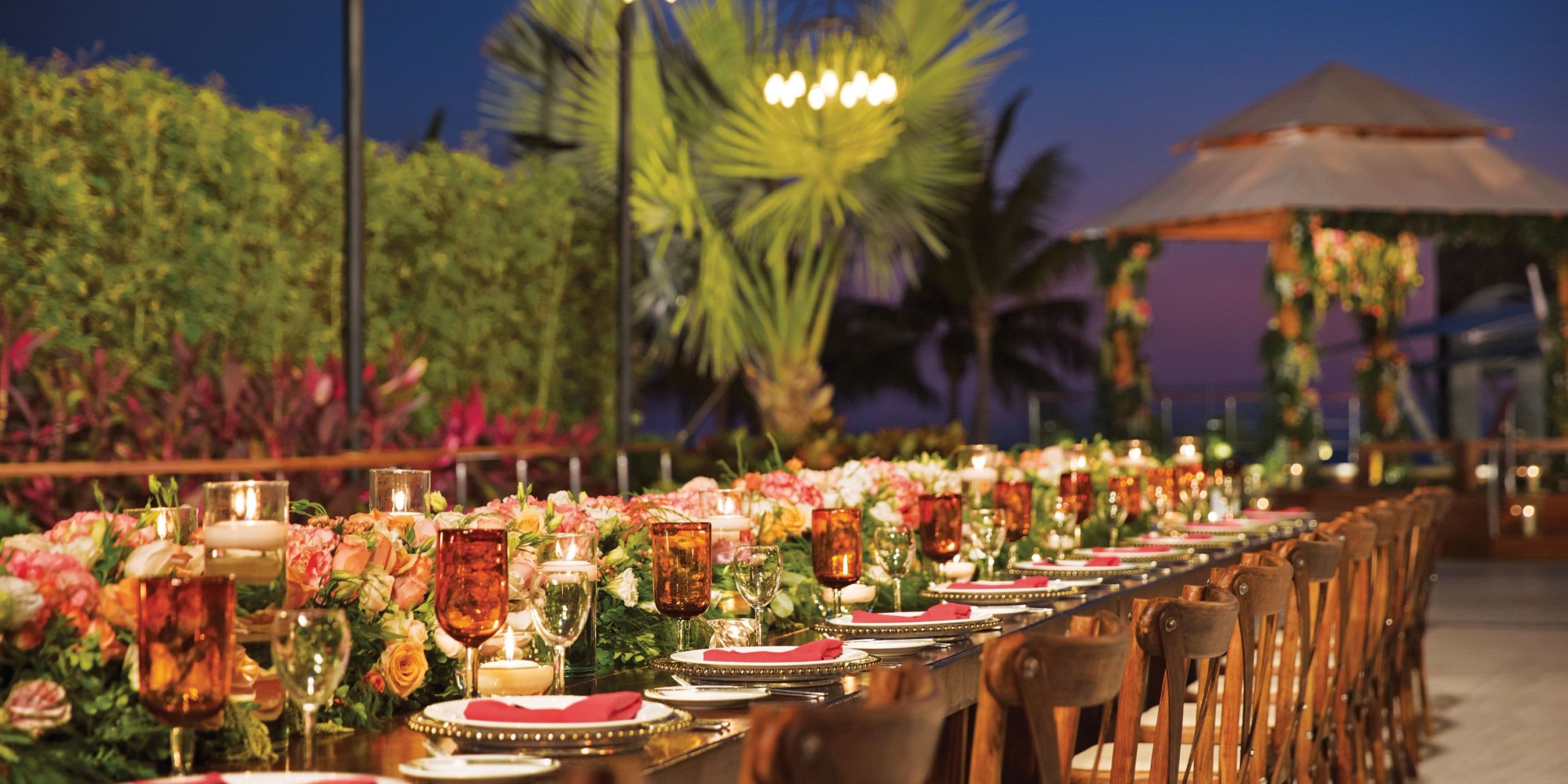 Create an enchanting outdoor dining experience at Dreams Vallarta Bay, where a long, elegantly decorated table showcases stunning floral arrangements, glowing candles, and sophisticated place settings. The presence of palm trees and gentle lighting enhances the ambiance with a tropical evening charm.