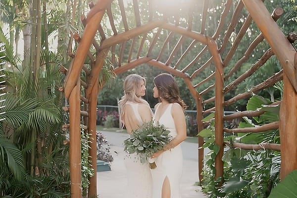 Two women in white dresses stand under a wooden arch at a destination wedding in El Dorado Casitas Royale.