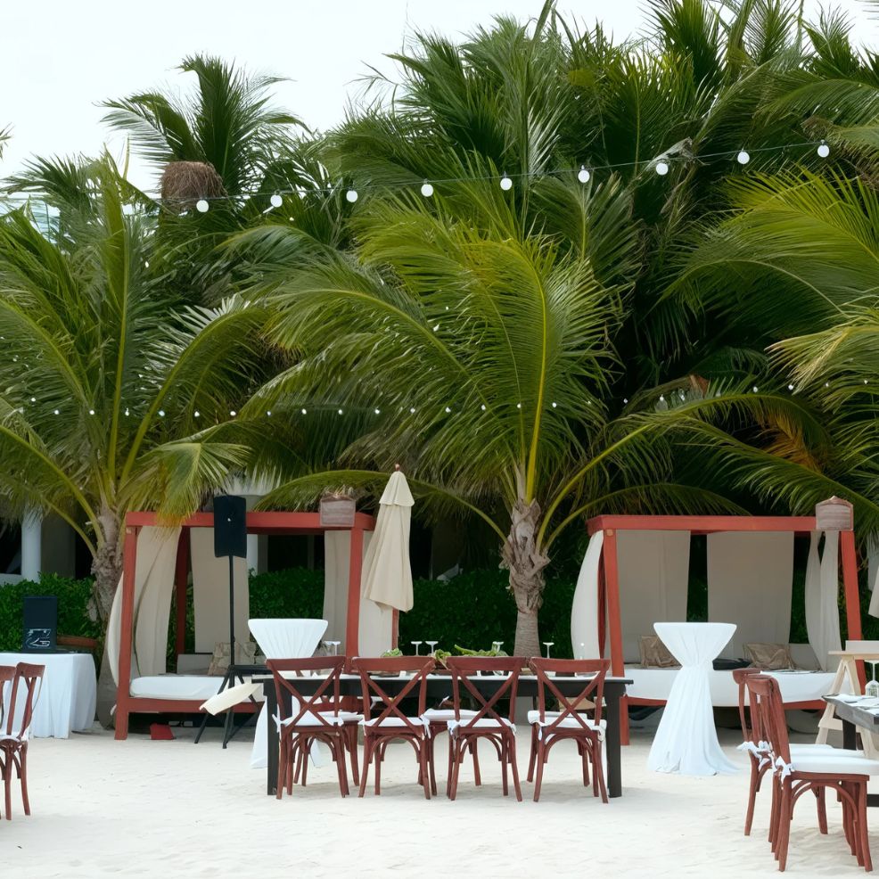 Outdoor beach setup with wooden chairs and tables, surrounded by cabanas and palm trees. String lights hang above the area.