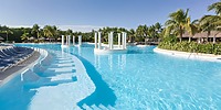 The Grand Palladium Kantenah features a spacious outdoor swimming pool with pristine blue water, encircled by palm trees and sun loungers. Floating columns grace the center, while huts are nestled in the background beneath a clear sky.