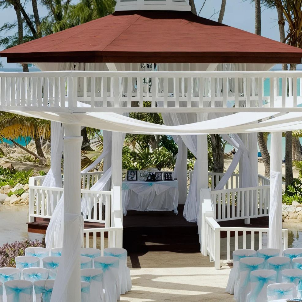 A white gazebo decorated with white drapes and blue chair sashes, set up for a wedding ceremony by the beach with palm trees in the background.