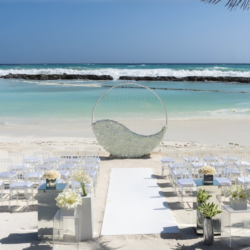 Destination wedding setup on the beach with white chairs, floral accents, and a modern arch by the sea.