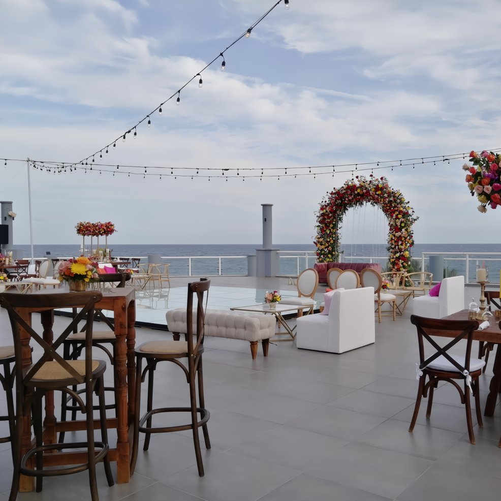 Destination wedding rooftop setup with tables, chairs, floral arch, and string lights above the ocean.