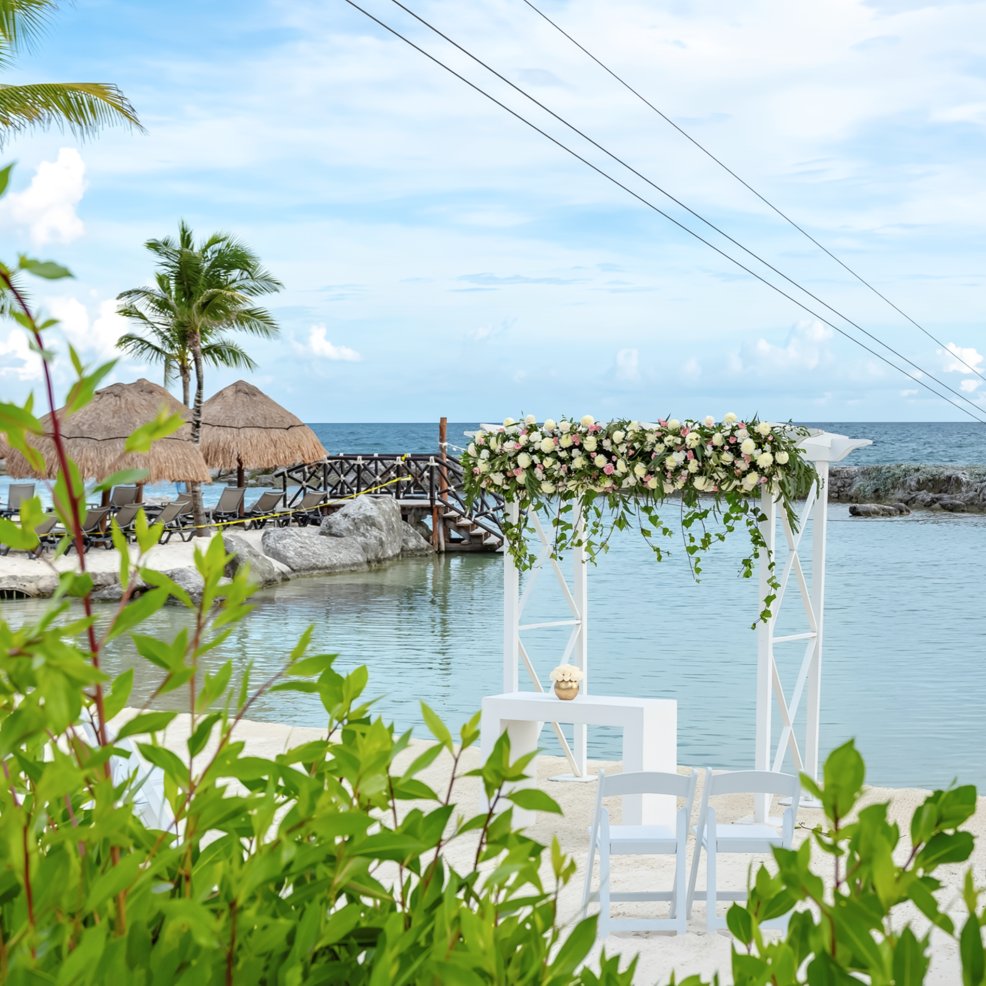 A destination wedding setup with a white altar and chairs arranged by the waterfront on a tropical beach.