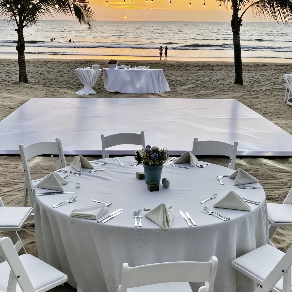 A beautifully set round table with white linens awaits guests on the beach, overlooking a dance floor and the ocean at sunset—the perfect setting for an unforgettable destination wedding.