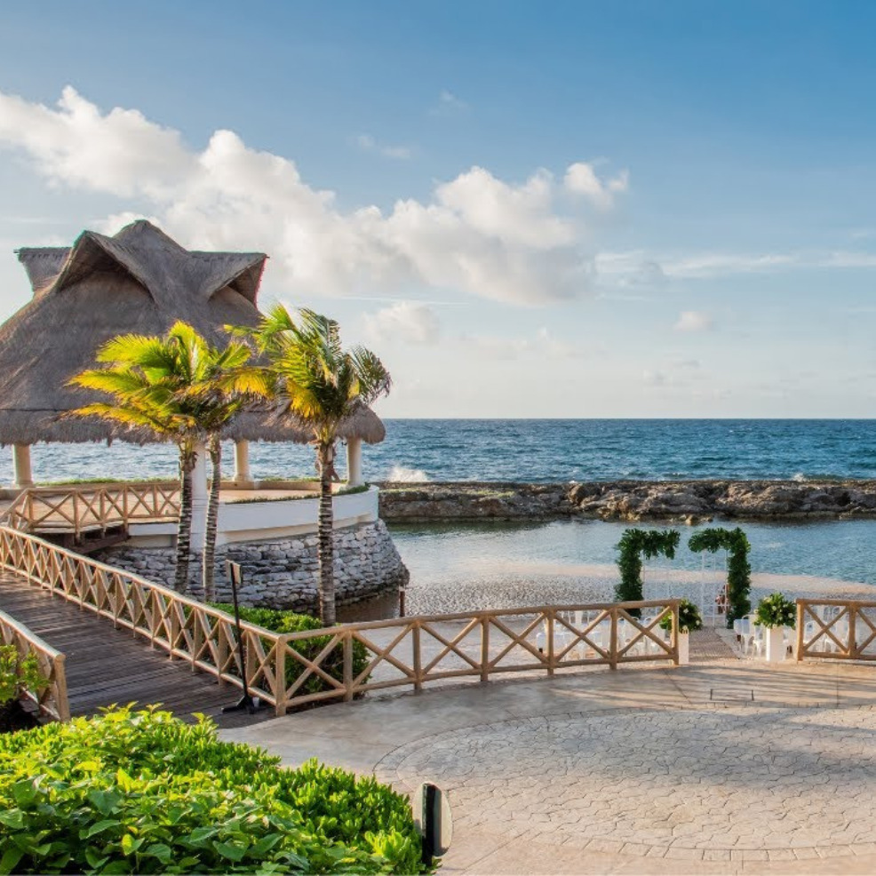 Destination wedding venue at Hard Rock Riviera Maya with a thatched gazebo, walkway, and stone altar by the sea, set under a clear blue sky.