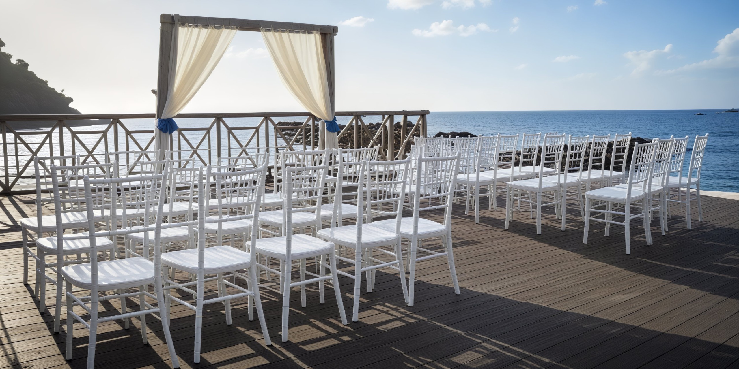 White chairs are elegantly arranged on a wooden deck for a picturesque seaside destination wedding ceremony.