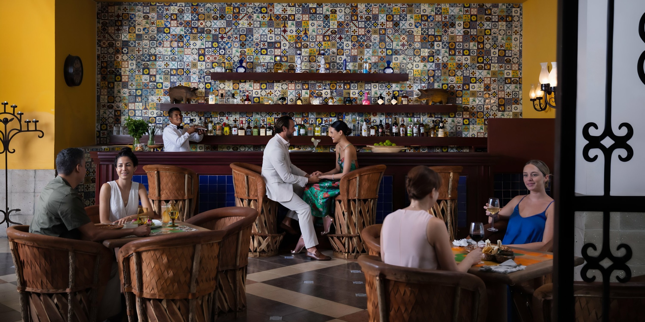 A vibrant bar scene at Hyatt Zilara Cancun with multiple patrons seated, a couple engaged in conversation at the bar, surrounded by colorful tiles and elegant decor.