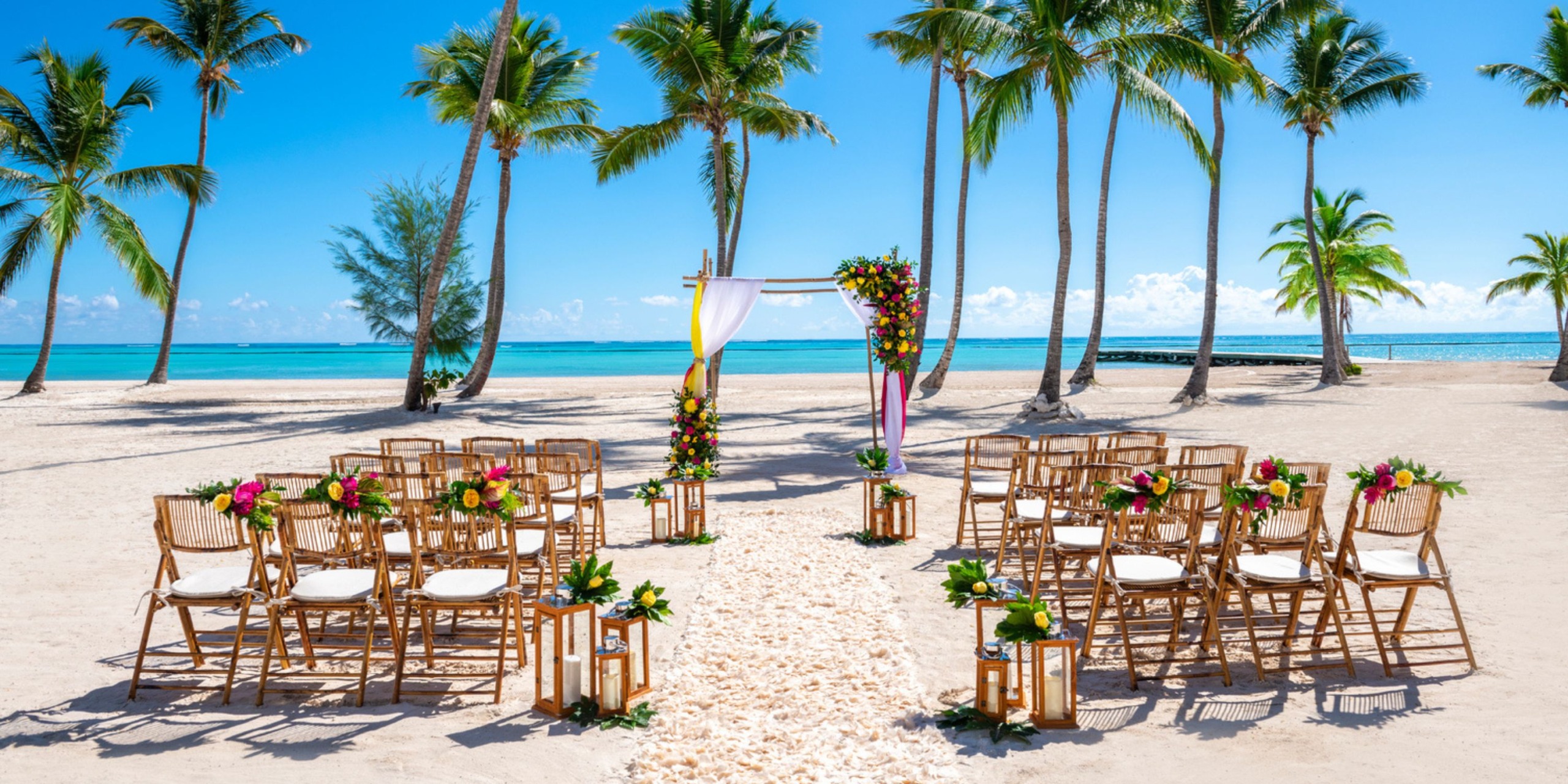 At the Hyatt Zilara Cap Cana, a destination wedding takes place on the beach, featuring chairs, a floral arch, and palm trees beneath a clear sky.