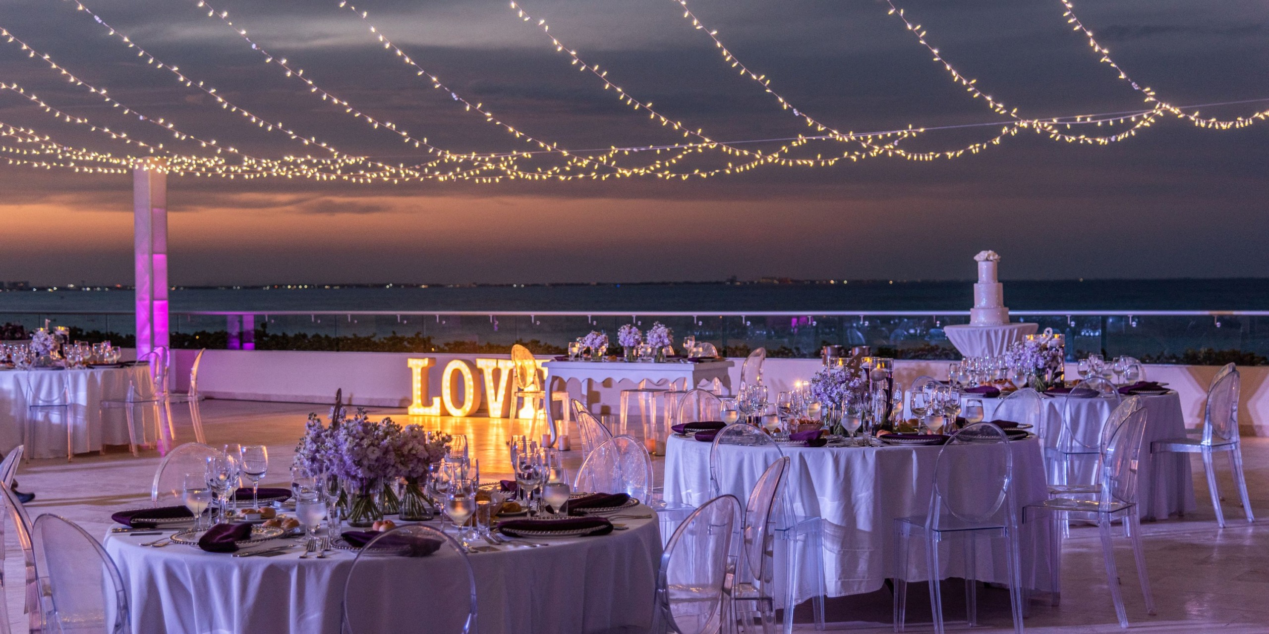 Experience an outdoor wedding reception at Hyatt Ziva Riviera Cancun, featuring round tables and transparent chairs, a "LOVE" sign illuminated by string lights overhead, all set against a breathtaking ocean view at sunset.