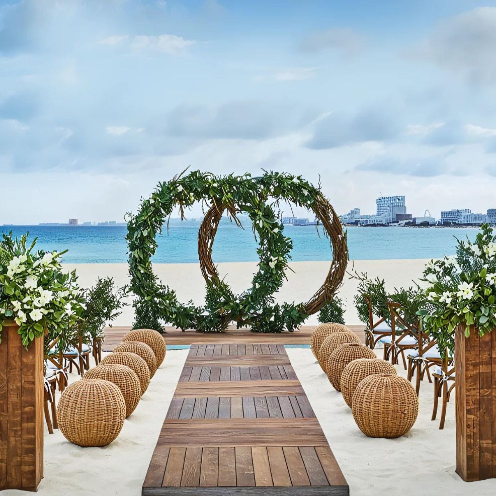 Outdoor beach wedding setup with a wooden aisle, circular floral arch, wicker seats, and greenery accents, overlooking the ocean and distant buildings.