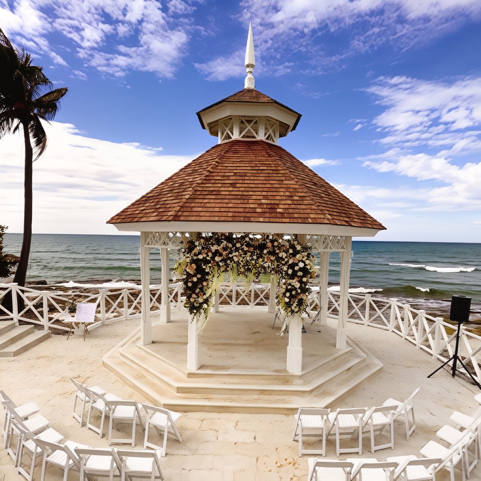 White gazebo adorned with flowers and wedding chairs, set by the ocean for a sunny destination wedding.