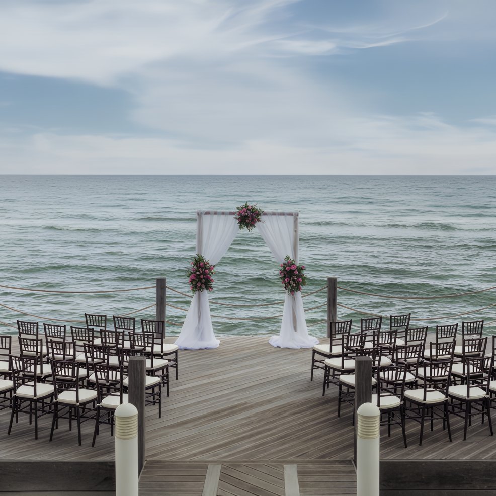 Destination wedding setup on a wooden deck by the ocean, featuring chairs and a floral arch.