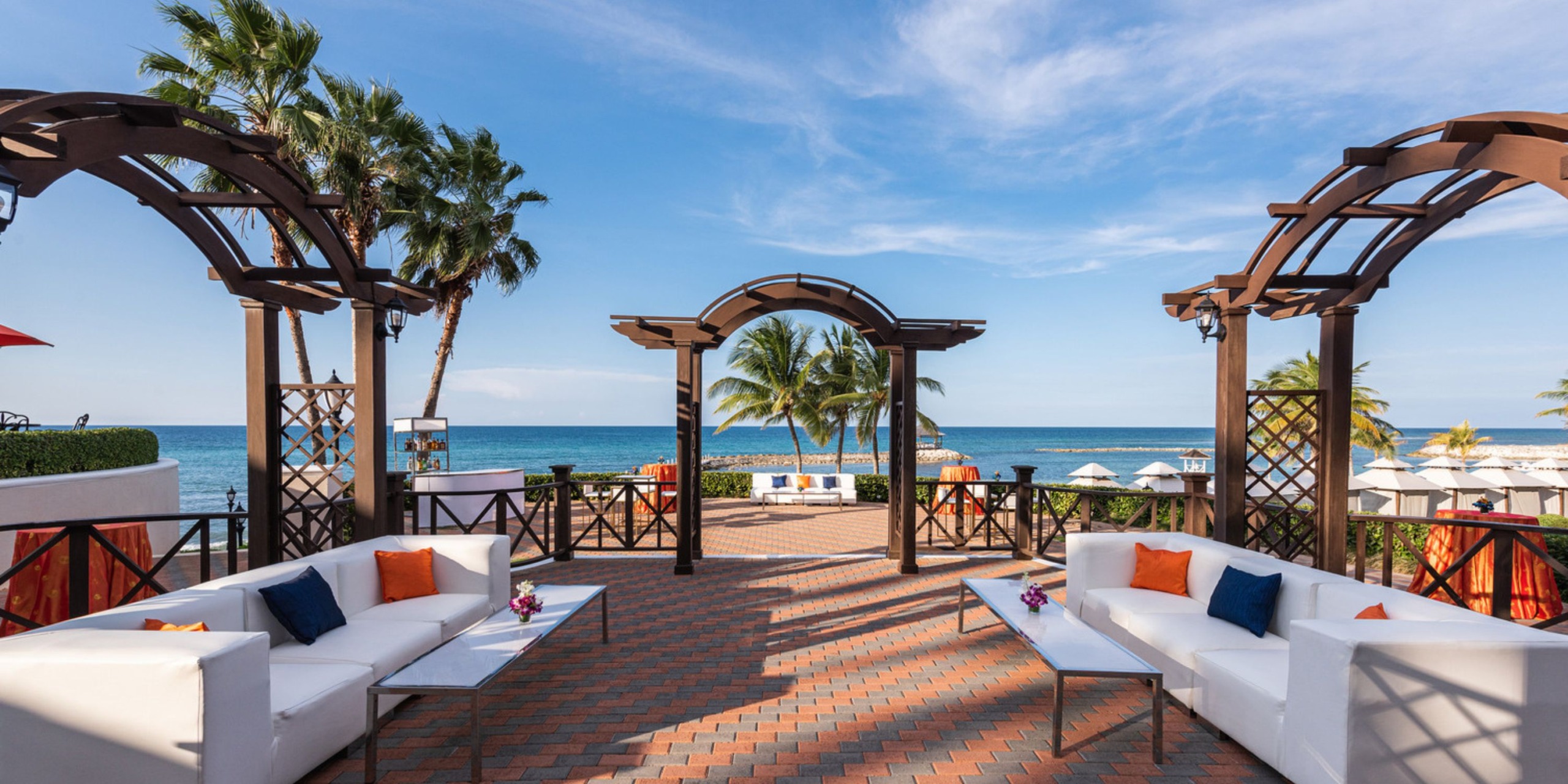 The Jewel Grande terrace, adorned with white sofas, offers a breathtaking ocean view, complemented by palm trees and elegant arches under a vibrant blue sky—an idyllic setting for a destination wedding.