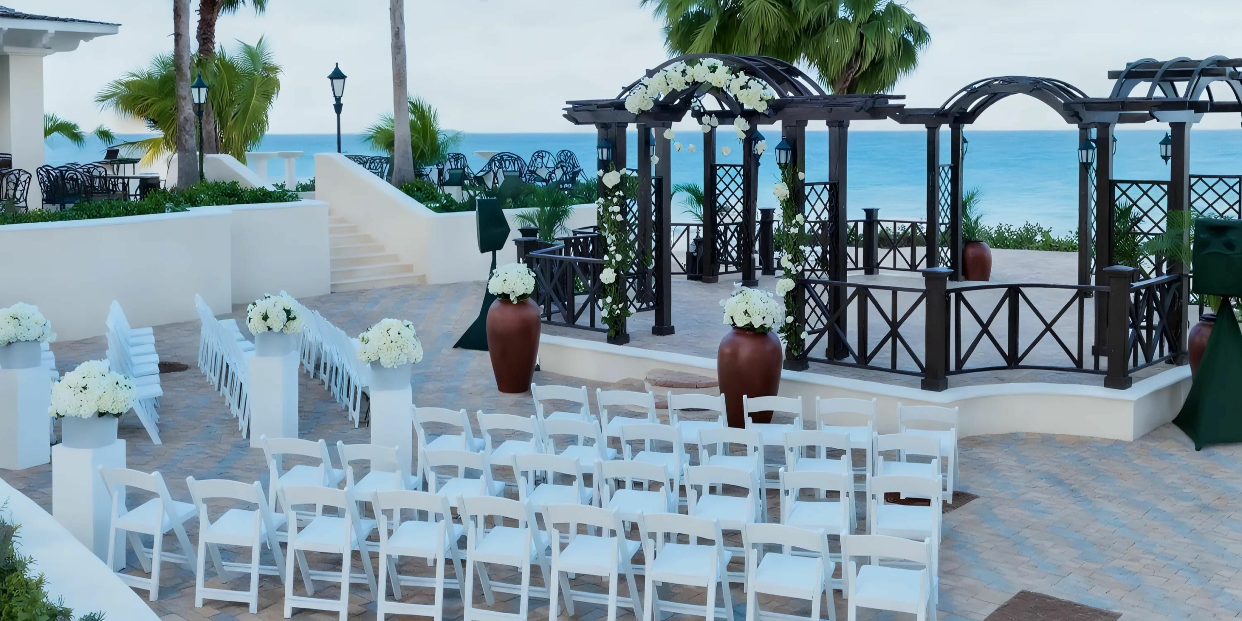 Outdoor wedding ceremony setup with white chairs arranged in rows facing a gazebo adorned with flowers, overlooking the ocean on a paved patio.