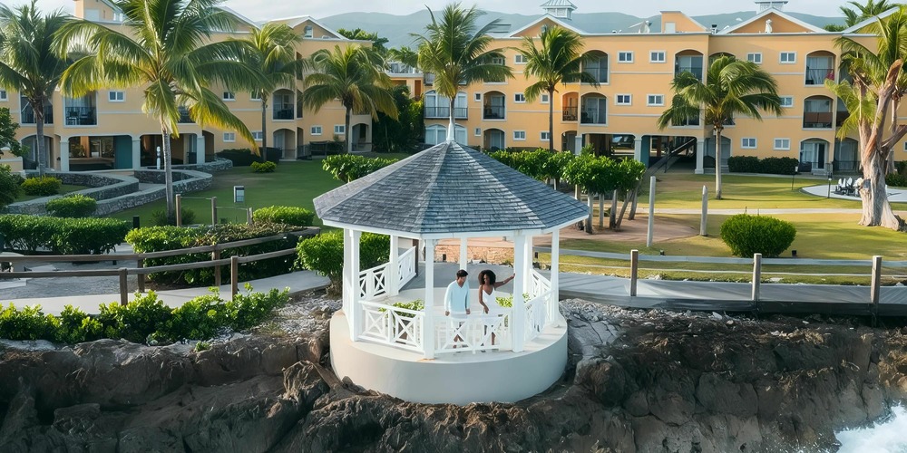 A white gazebo overlooks a rocky shore with people inside, surrounded by palm trees and facing a yellow resort building.