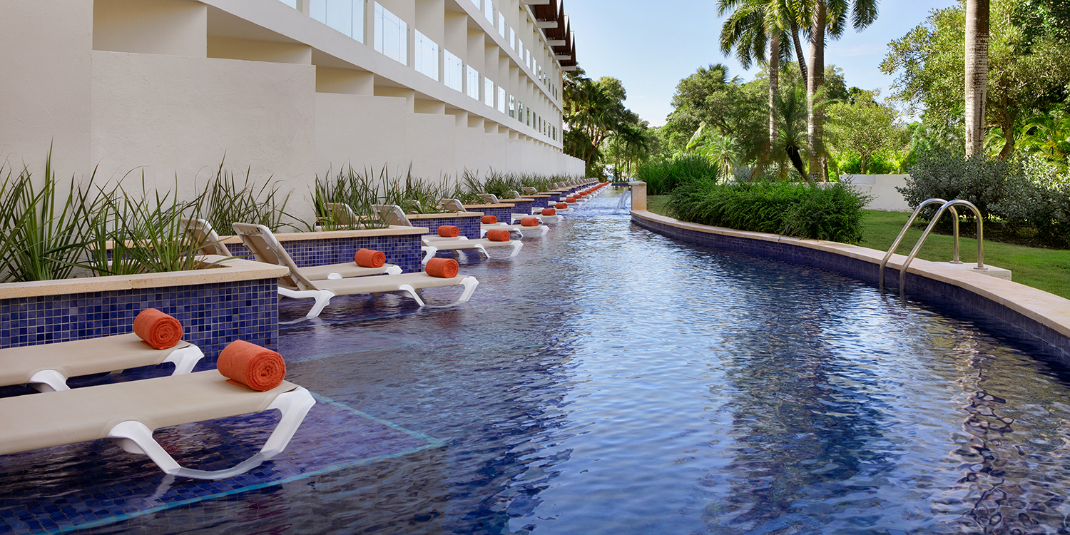A serene Hilton La Romana pool lined with loungers and vibrant orange towels, surrounded by lush greenery under a clear sky, perfect for destination weddings.