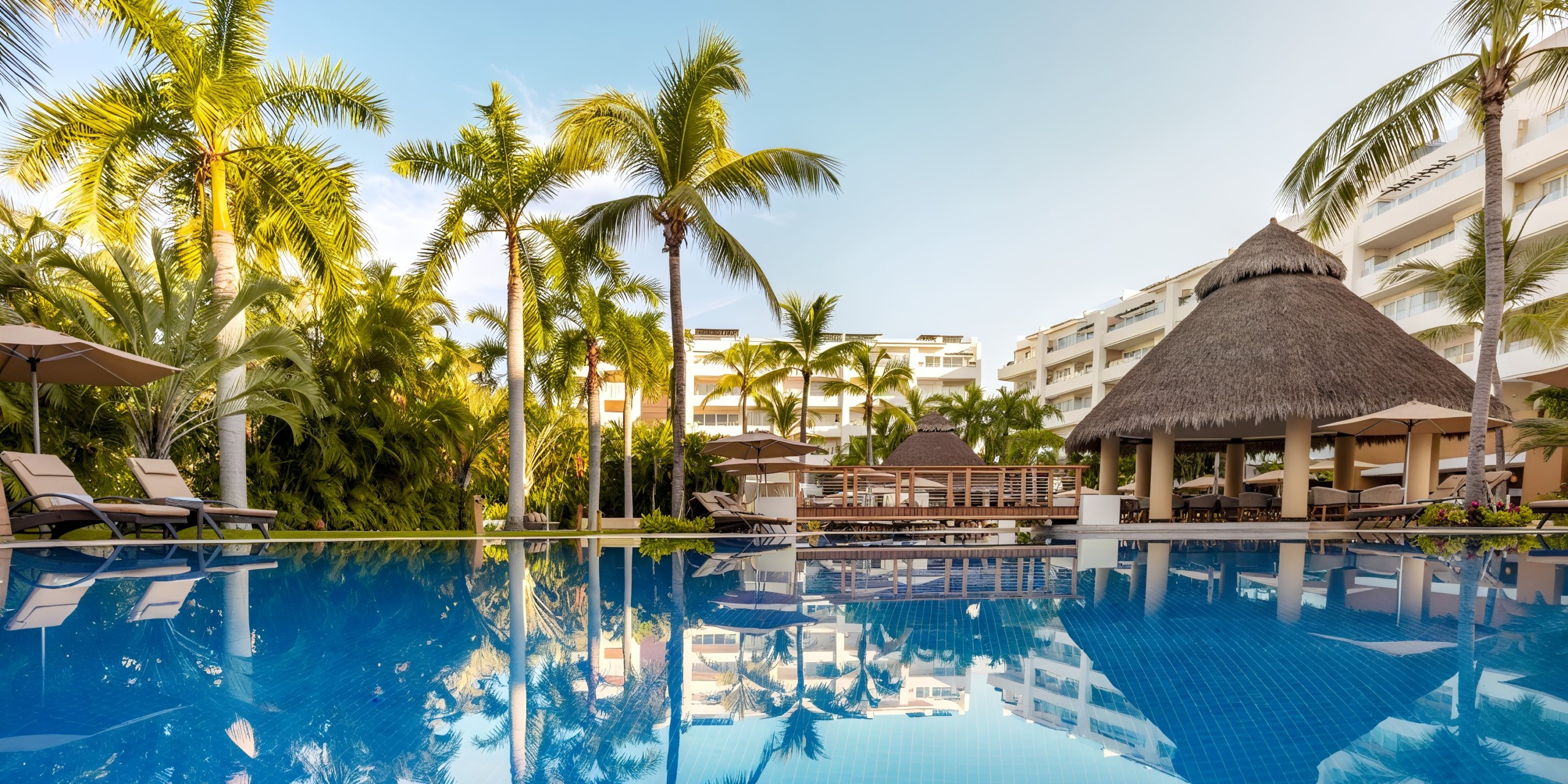The Marival Distinct features a swimming pool encircled by palm trees, loungers, and a thatched-roof hut, with the hotel's elegant structure set against a backdrop of clear blue skies.