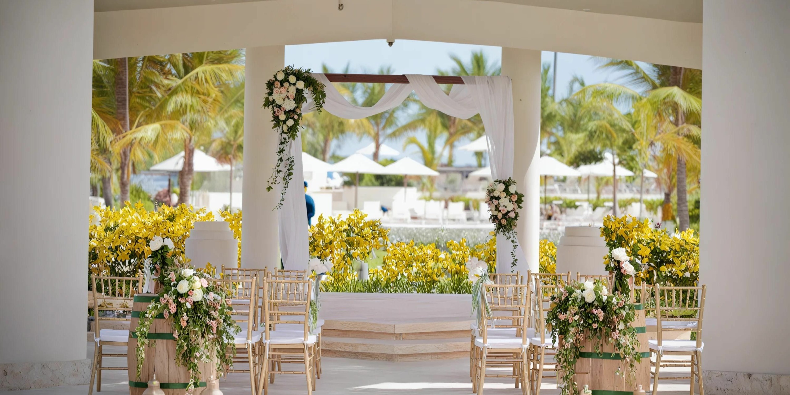 A Dreams Onyx wedding altar adorned with white drapery and floral arrangements is set up outdoors. Bamboo chairs featuring floral decorations are meticulously arranged in rows, while tropical plants create a lush, surrounding backdrop.