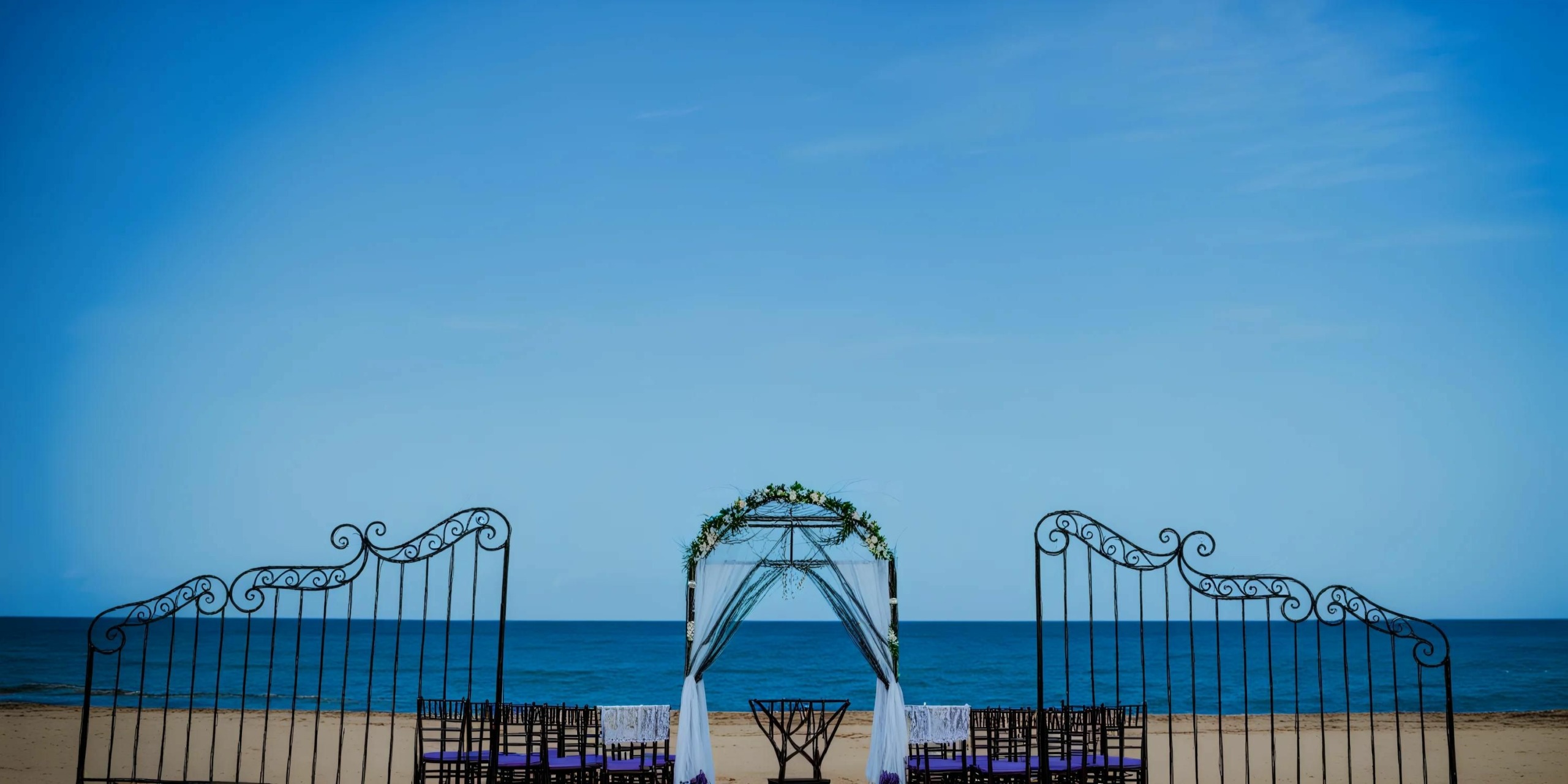 A Dreams Onyx beach wedding setup features a white arch adorned with greenery, flanked by black decorative gates, all facing the calm ocean under a clear blue sky.