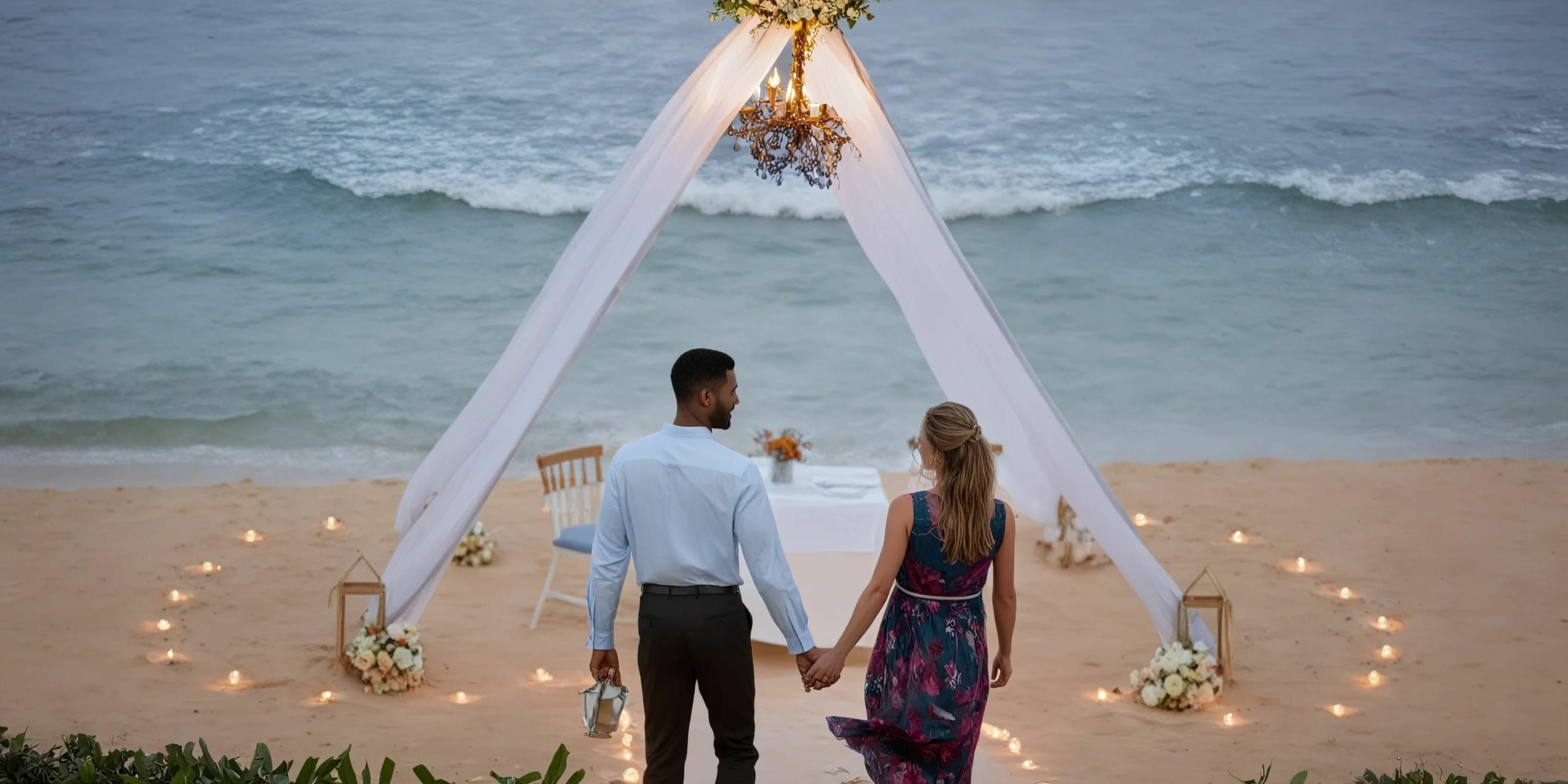 A couple holding hands walks toward a decorated canopy on the beach at Hilton Playa del Carmen. A chandelier and flowers adorn the canopy, with chairs and a table set behind it.
